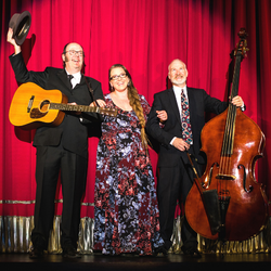 Image of The Beggars, a musical trio, two males standing either side of a female lead singer. One male thrusts his hat in the air holding a guitar while the other male holds a double bass. They are standing in front of a red stage curtain.