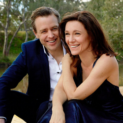 A man dressed in a suit with a white shirt and open collar, sitting next to a woman, wearing a navy blue silk gown. Both are smiling.
