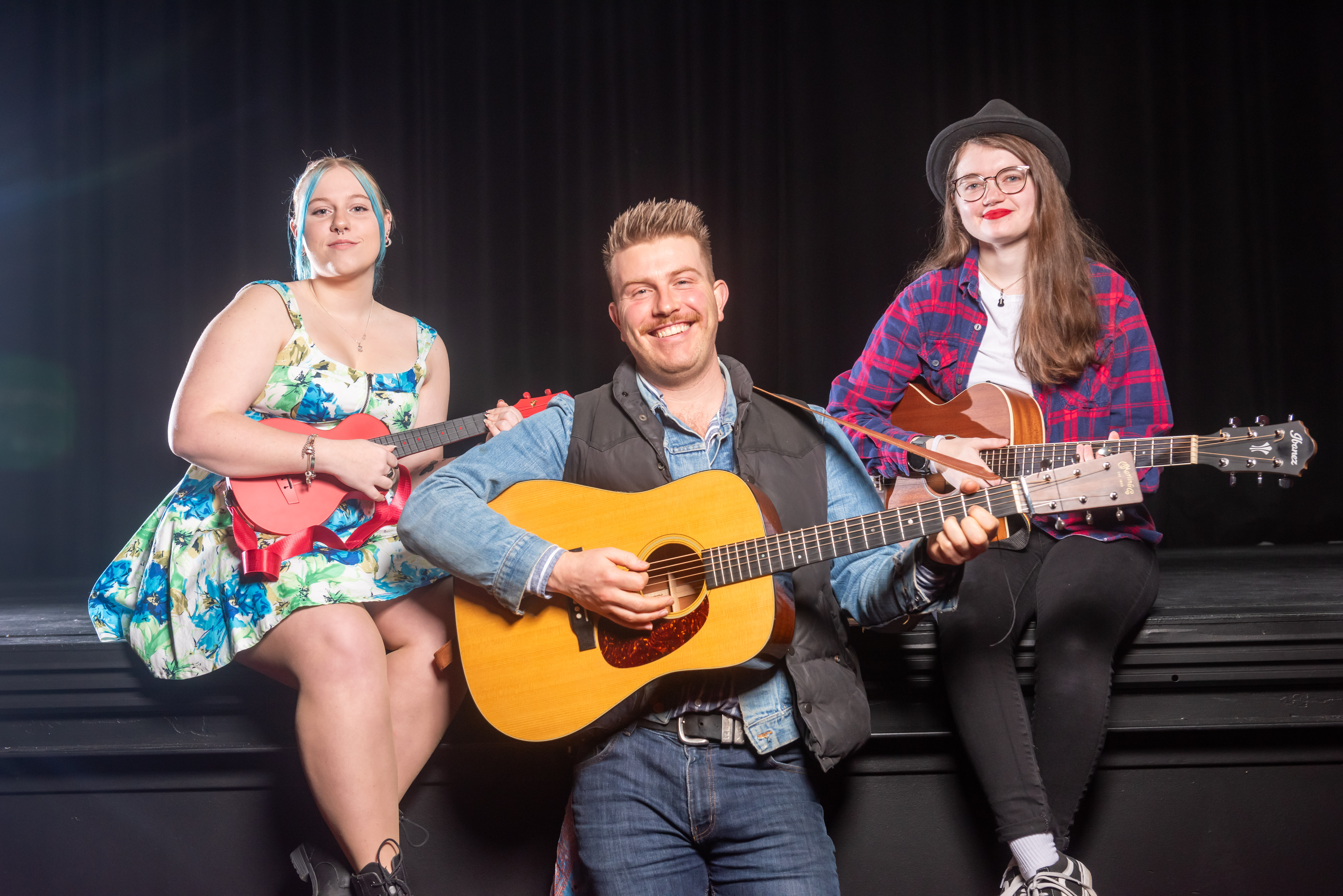 Two musicians sitting on a stage with guitars and one standing in front of the stage with a guitar