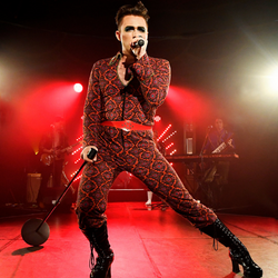 A man wearing a full red and black diamond patterned jumpsuit, is on stage with red lighting, holding a microphone on a stand, singing.