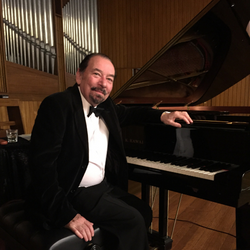 A mature man sitting at a piano with its lid open, resting his arm on the top, dressed in a tuxedo.