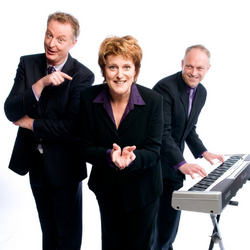 Three people, two males and a female, dressed in black suits, smiling. One is playing the keyboard.