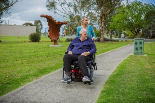 A lady pushing a man in a wheelchair along a walking path. 